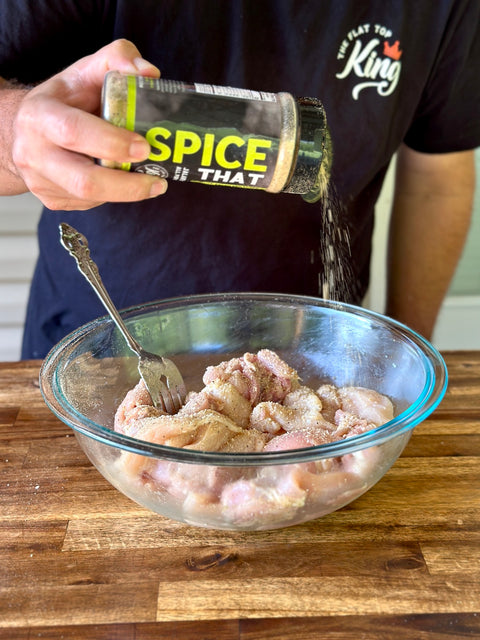 Person seasoning chicken in a glass bowl with 'SPICE THAT' container.